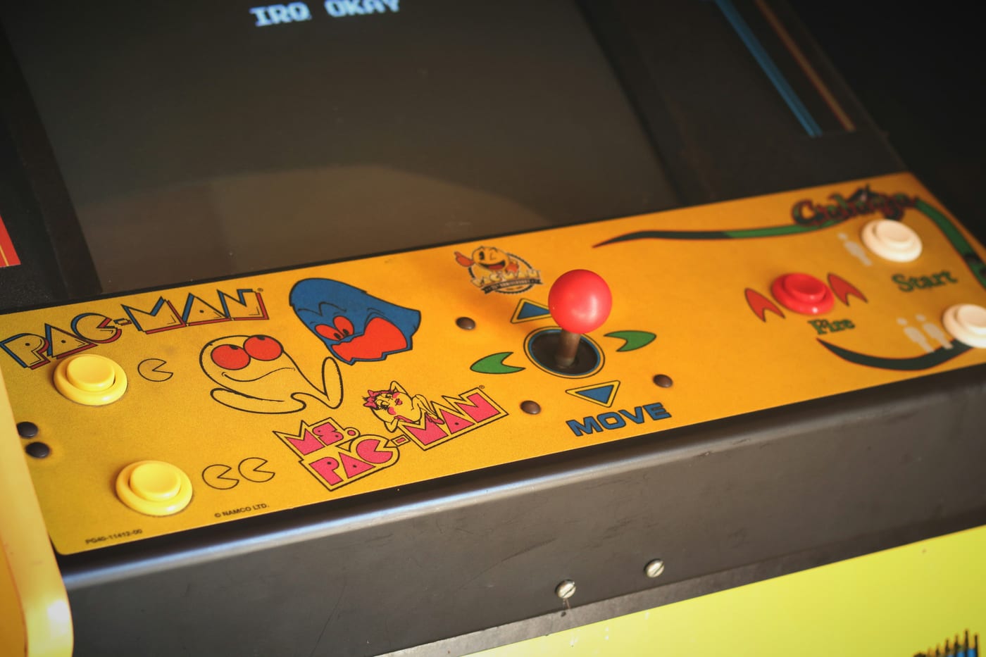 A worn Pac-Man and Ms. Pac-Man arcade control panel, yellow paint chipped from thousands of quarters, a single joystick labeled MOVE — a metaphor for chasing a score that means nothing outside the cabinet