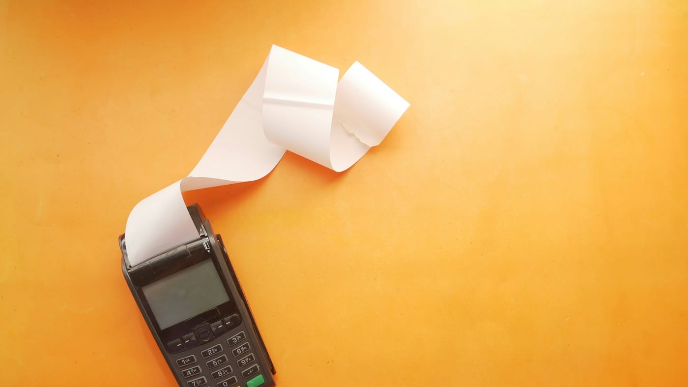 A payment terminal on an orange surface with a long paper receipt curling out of it, a metaphor for a token bill that keeps printing