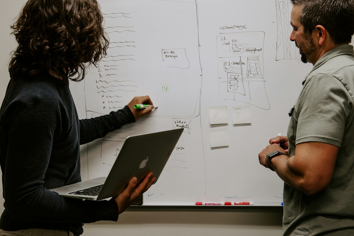 Two people at a whiteboard covered in sketches, one pointing at the design while the other listens, a metaphor for the review call where a candidate defends their own decisions