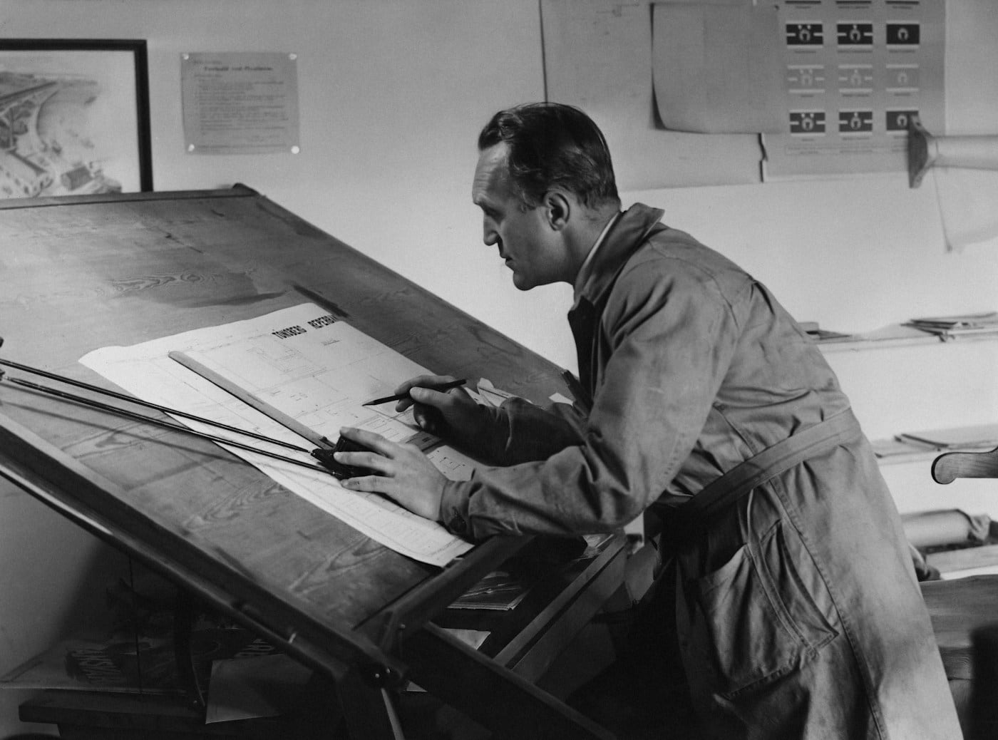 An engineer in a work coat bent over a drafting board with a pencil and ruler, a metaphor for thinking a design through alone, at your own pace