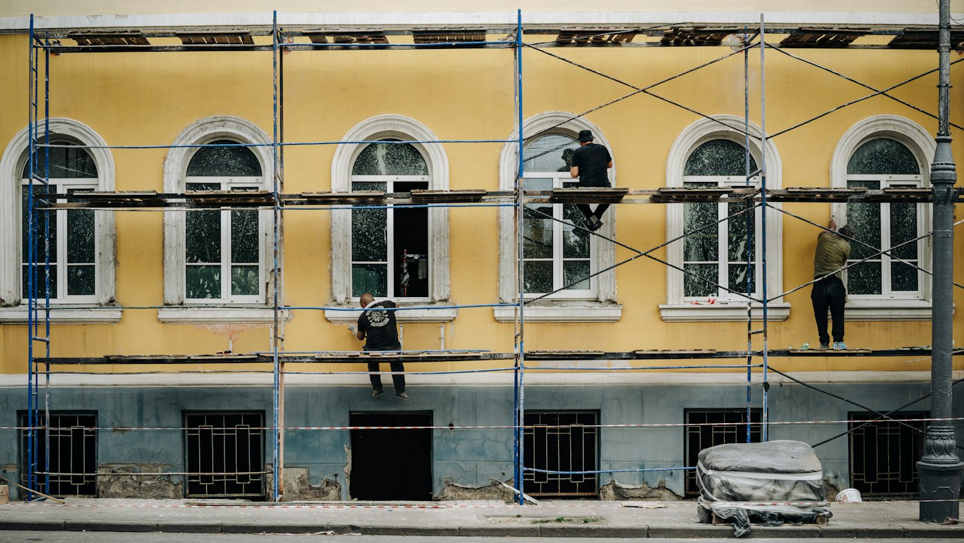 Workers on metal scaffolding in front of a finished yellow building facade, a metaphor for agent architectures built around a model that can now stand on its own